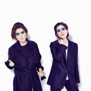 Sara in a black overcoat with vibrant green eye makeup, and Tegan in a black suit jacket with vibrant blue eye makeup.