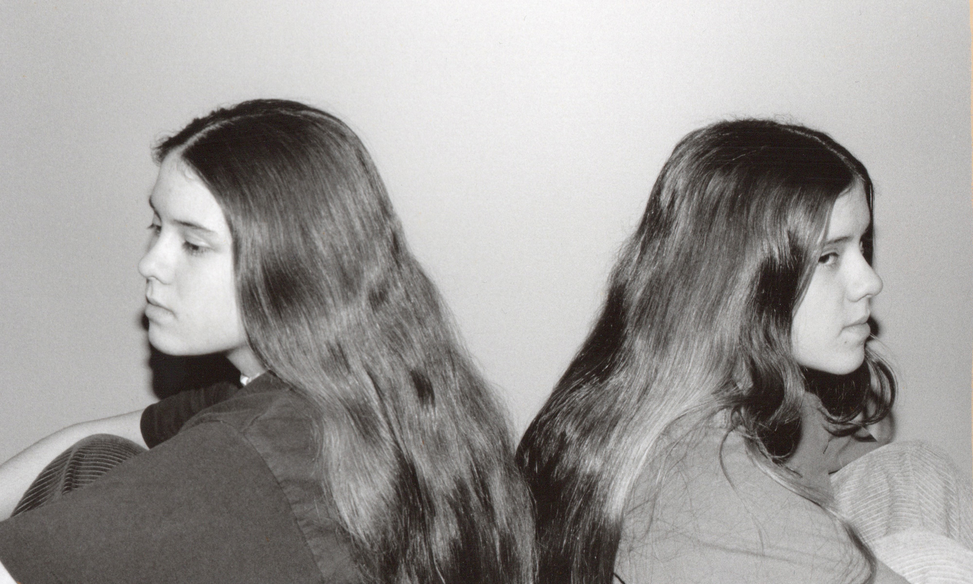 Black and white photo of high school Tegan and Sara with long hair, sitting back-to-back