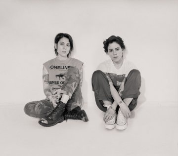 Black and white long shot of Tegan and Sara sitting in a photo studio setting on a white background