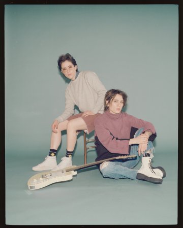 Long shot in a photo studio setting, with Sara sitting on a stool and Tegan on the floor with a guitar